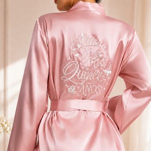 May include: A light pink satin robe with long sleeves and a matching belt. The back of the robe features white embroidered text and a crown design. The text reads "Mis Quince Años".