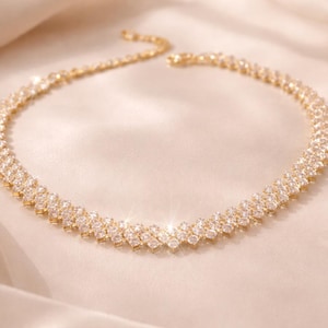 May include: A gold-toned necklace with a double-row design, encrusted with sparkling clear crystals. The necklace is displayed on a satin surface, reflecting light. The jewelry piece is a statement accessory.