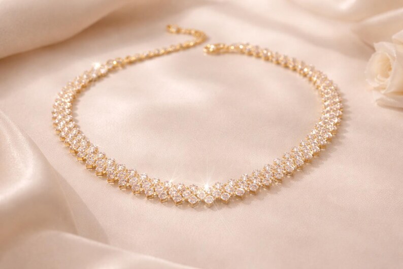 May include: A gold-toned necklace with a double-row design, encrusted with sparkling clear crystals. The necklace is displayed on a satin surface, reflecting light. The jewelry piece is a statement accessory.