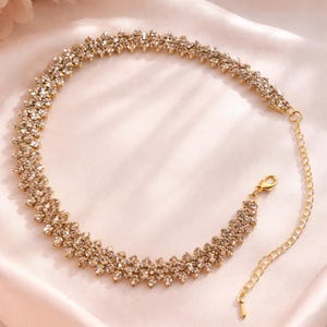 May include: A gold-toned necklace with a floral design, set with numerous small, sparkling crystals. The necklace is displayed on a soft, pink satin surface. The necklace has a delicate chain and clasp.