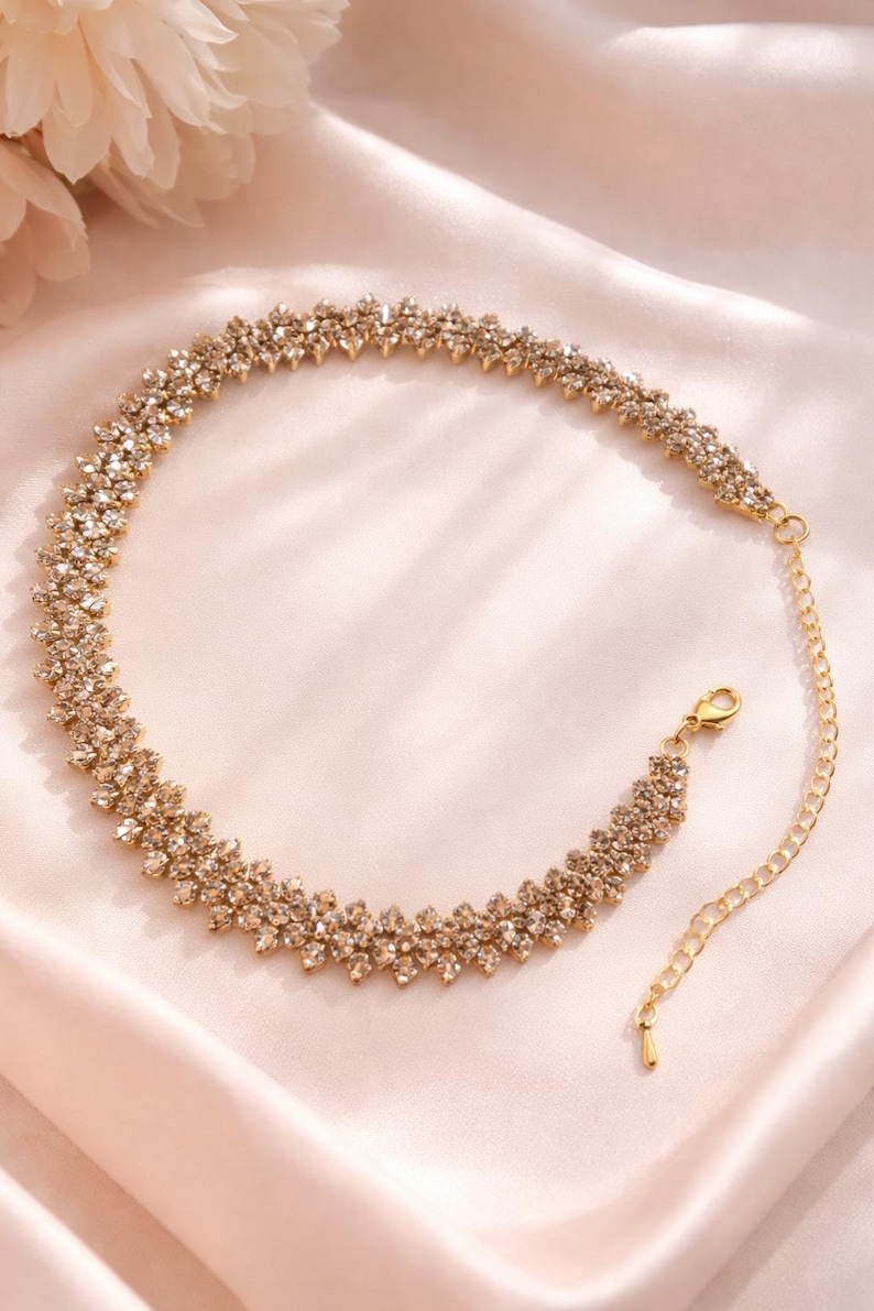 May include: A gold-toned necklace with a floral design, set with numerous small, sparkling crystals. The necklace is displayed on a soft, pink satin surface. The necklace has a delicate chain and clasp.