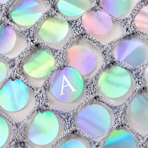 May include: A gray crocheted net fabric featuring circular iridescent sequins attached within each mesh opening. One of the circular sequins prominently displays the capital letter A in a white serif font, contrasting with the shimmering surface.