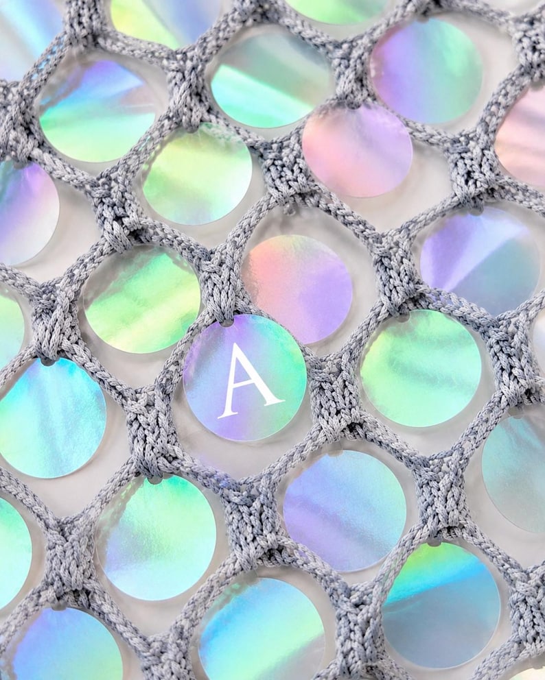 May include: A gray crocheted net fabric featuring circular iridescent sequins attached within each mesh opening. One of the circular sequins prominently displays the capital letter A in a white serif font, contrasting with the shimmering surface.