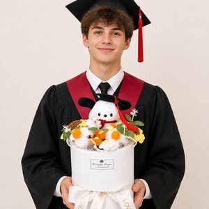 Może przedstawiać: Prezent na ukończenie studi&oacute;w. Białe, okrągłe pudełko zawiera kompozycję kwiatową i pluszową zabawkę w czapce absolwenta. Pudełko trzyma osoba w todze i czapce absolwenta. Pudełko ma wstążkę z napisem "LOVE IS LOVE".
