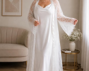 May include: A white floor-length satin slip dress with a cowl neckline paired with a matching long-sleeved white lace robe featuring wide sleeves and scalloped lace edges. The ensemble is completed with white fluffy open-toe slide sandals.