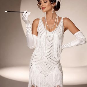 May include: A white sleeveless dress featuring silver sequined chevron patterns and long fringe detailing. The ensemble includes white satin elbow-length gloves, a multi-strand pearl necklace, a silver headpiece with white feathers, and a black cigarette holder.