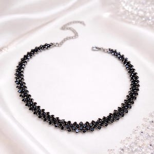 May include: A black beaded necklace with a zig-zag design. The necklace is made of small, dark beads and has a silver-colored chain clasp. The necklace is displayed on a white, silky surface.