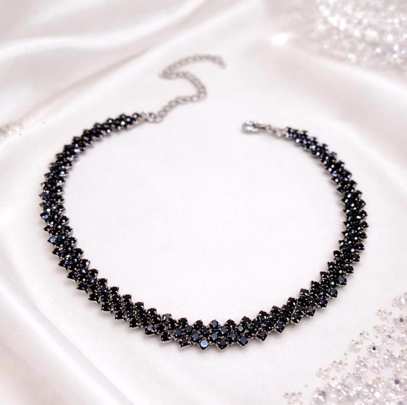 May include: A black beaded necklace with a zig-zag design. The necklace is made of small, dark beads and has a silver-colored chain clasp. The necklace is displayed on a white, silky surface.