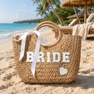 May include: A tan woven straw tote bag featuring the white text BRIDE in block letters with gold trim. A white ribbon is tied around one of the two rounded handles. A small white heart patch is positioned below the text on the front panel.