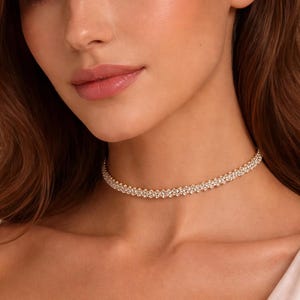 May include: A close-up of a woman wearing a delicate, sparkling choker necklace. The necklace is made of small, round, clear crystals set closely together, creating a continuous band of shimmer. The necklace sits around the base of the neck.