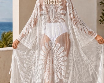 May include: A sheer white lace beach cover-up featuring an intricate floral pattern and wide sleeves. The back of the garment displays the name NICOLE in bold, capitalized letters. The fabric is lightweight and semi-transparent with scalloped edges.