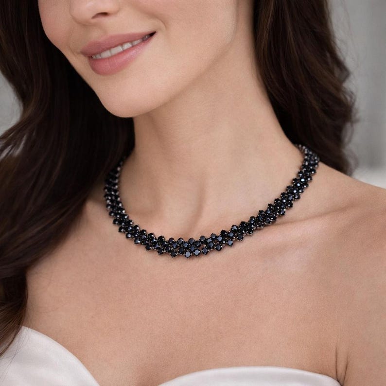 May include: A black beaded necklace with a multi-strand design. The necklace is made of small, dark beads and sits close to the neckline. The jewelry is worn by a person.