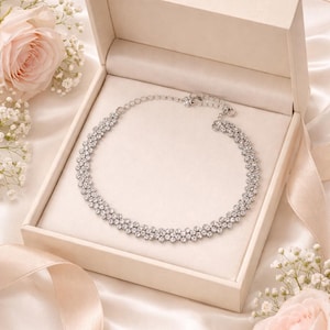 May include: A silver necklace with multiple rows of sparkling crystals rests in an open, cream-colored box. The box sits on a satin surface, with a ribbon and pink roses in the background. The necklace is a statement piece.