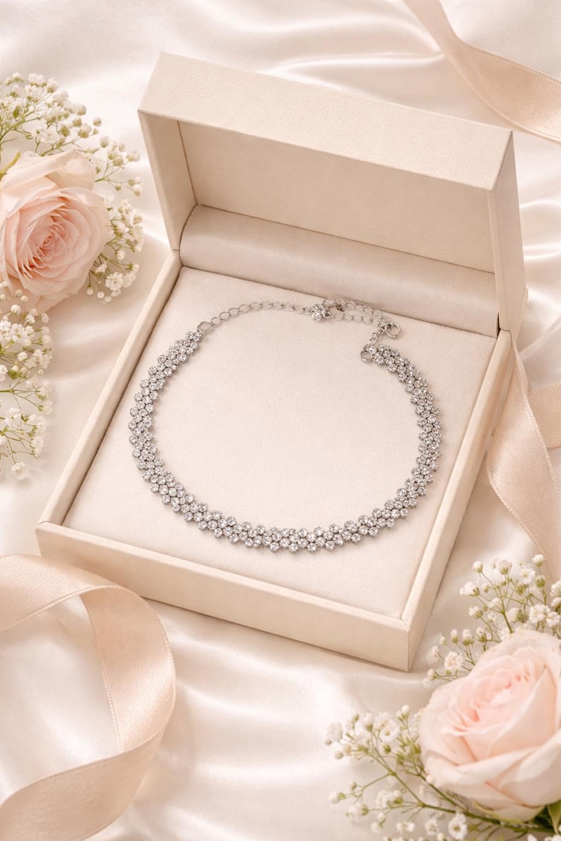 May include: A silver necklace with multiple rows of sparkling crystals rests in an open, cream-colored box. The box sits on a satin surface, with a ribbon and pink roses in the background. The necklace is a statement piece.