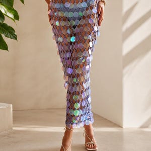 May include: A floor-length lavender crochet skirt featuring an open-knit mesh pattern adorned with numerous circular, iridescent, light-reflecting sequins throughout the entire garment, creating a shimmering effect against the light purple yarn base.