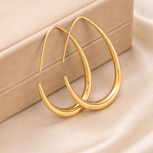 May include: A pair of gold-colored teardrop hoop earrings. The earrings have a smooth, polished finish and a simple, elegant design. They are displayed on a neutral-toned surface, suggesting a jewelry or fashion theme.