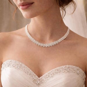 May include: A sparkling, silver-toned necklace with a delicate, layered design. The necklace is worn with a white, strapless wedding dress, featuring intricate beadwork along the neckline. The image is a close-up, focusing on the jewelry and dress details.