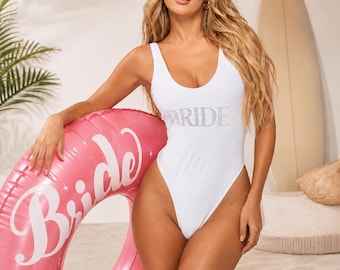 May include: A white one-piece swimsuit featuring the word BRIDE in silver rhinestones on the chest. The swimsuit has a scoop neckline and a high-cut leg design. Next to it is a large, round, pink inflatable pool float with the word Bride.
