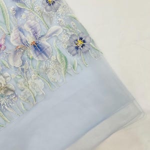 Puede incluir: Una tela transparente de color azul claro con intrincados bordados florales en tonos morado, blanco y verde. El diseño incluye flores de iris detalladas y flores pequeñas acentuadas con lentejuelas y cuentas transparentes.