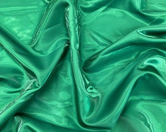May include: Emerald green satin fabric with a glossy sheen. The fabric is draped, creating folds and shadows that highlight its texture. The material appears smooth and flowing, suitable for apparel or decorative projects.