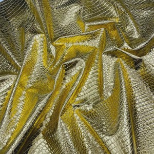 May include: A close-up shot of a golden, textured fabric with a crocodile or alligator skin pattern. The material is shiny and reflective, with folds and creases that create shadows and highlights. The overall effect is luxurious and opulent.