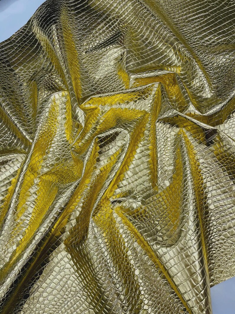 May include: A close-up shot of a golden, textured fabric with a crocodile or alligator skin pattern. The material is shiny and reflective, with folds and creases that create shadows and highlights. The overall effect is luxurious and opulent.