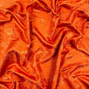 Op de afbeelding: Fel oranje stof met een bloemenpatroon. De stof heeft een satijnen glans en is gedrapeerd, waardoor schaduwen en highlights ontstaan. Het bloemenpatroon is subtiel in reliëf, wat textuur toevoegt. Geschikt voor kleding of interieur.