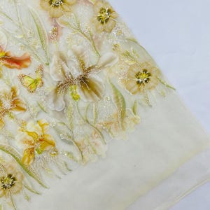 May include: A sheer off-white fabric featuring intricate floral embroidery in shades of yellow, gold, and green. The design includes detailed iris flowers, small leaves, and a butterfly, all embellished with clear sequins and tiny white beads.