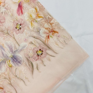 Puede incluir: Una tela transparente de color melocotón claro con intrincados bordados florales. El diseño incluye motivos de iris y flores pequeñas en tonos rosa, morado, amarillo y crema, acentuados con delicadas lentejuelas transparentes y cuentas.