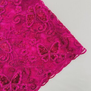 May include: Close-up of a vibrant fuchsia lace fabric. The fabric features embroidered butterflies and floral patterns, embellished with sequins. The edges are scalloped, adding a delicate touch to the design. The overall aesthetic is feminine and decorative.