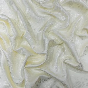 May include: Ivory-colored fabric with a subtle floral pattern. The fabric is draped, creating soft folds and shadows. The floral design is a tone-on-tone pattern, adding texture and visual interest. The fabric has a slight sheen, suggesting a satin or silk-like material.