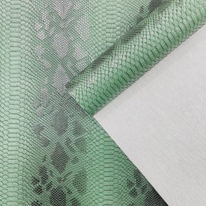 May include: Close-up of a green snakeskin pattern fabric. The fabric has a textured surface with a scale-like design in shades of green and grey. A portion of the fabric is rolled, revealing a white backing.