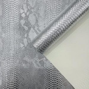 Puede incluir: Tela de cuero sintético plateada con un patrón de piel de serpiente. El material está enrollado en la esquina superior derecha, revelando un reverso blanco. La tela tiene una superficie brillante y texturizada.