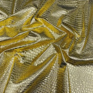 May include: A close-up shot of a gold-colored fabric with a crocodile skin texture. The fabric is folded and draped, creating shadows and highlights that emphasize the texture. The material has a shiny, metallic appearance.