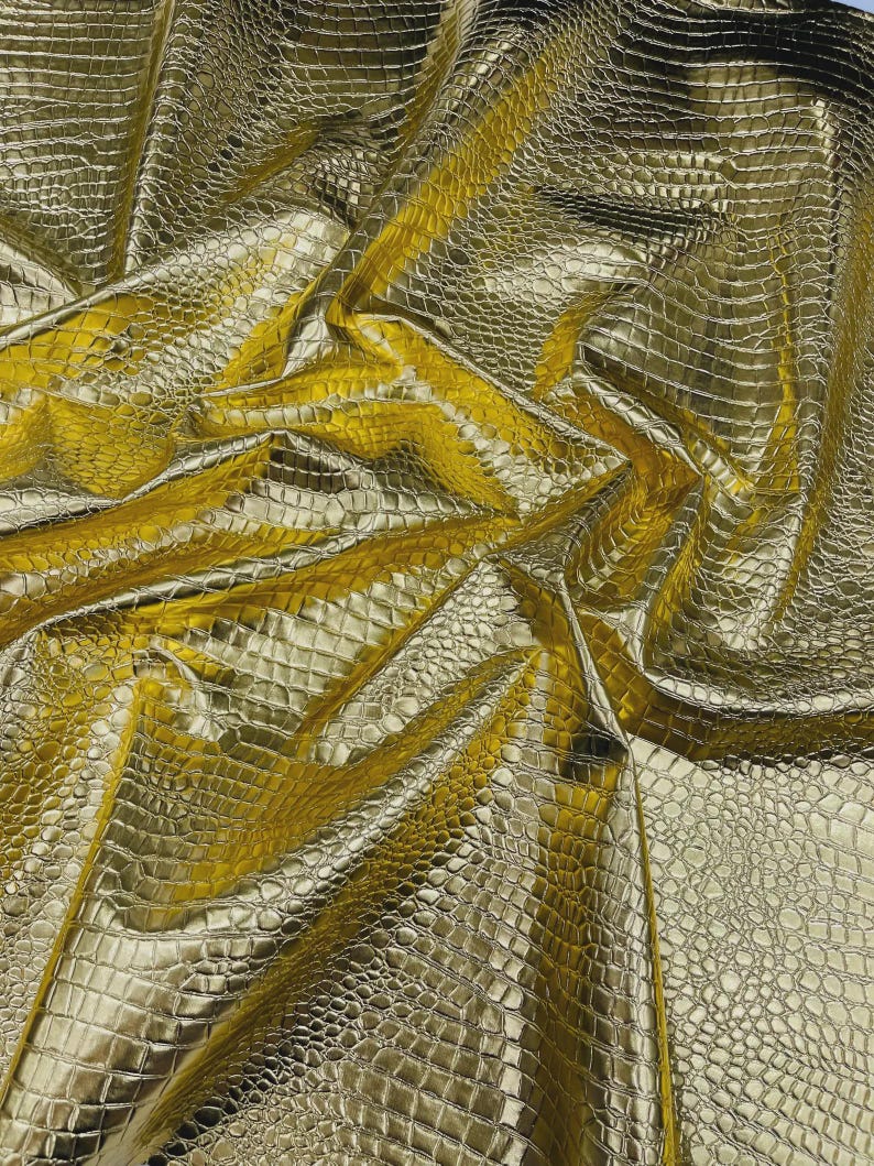 May include: A close-up shot of a gold-colored fabric with a crocodile skin texture. The fabric is folded and draped, creating shadows and highlights that emphasize the texture. The material has a shiny, metallic appearance.