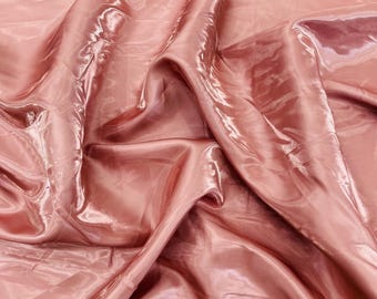 May include: A close-up shot of a flowing, shiny fabric in a soft, dusty rose color. The material has a silky, almost liquid appearance, with highlights and shadows creating depth. The fabric is draped and folded, showing its texture and sheen.