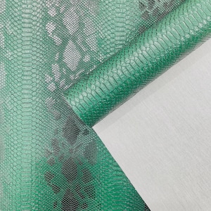 Op de afbeelding: Close-up van groene imitatie slangenleer stof met een getextureerd, schubachtig patroon. Het materiaal is opgerold en onthult een witte achterkant. De stof heeft een donker, gevlekt ontwerp en is geschikt voor knutsel- en designprojecten.