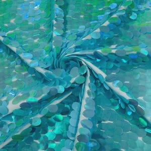 May include: Close-up of a fabric with a pattern of iridescent sequins in shades of teal and green. The sequins are arranged in a dense, overlapping pattern, creating a shimmering effect. The fabric is draped, revealing a white backing.