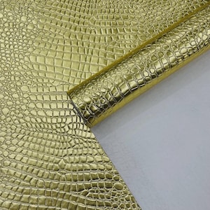May include: A close-up of a roll of gold-colored faux leather with a crocodile skin texture. The material is shiny and reflects light, with a detailed pattern of scales. The material is partially rolled, revealing a white backing.
