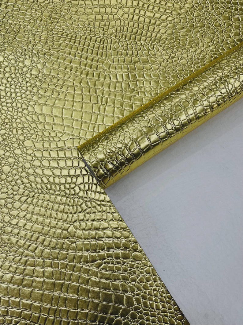 May include: A close-up of a roll of gold-colored faux leather with a crocodile skin texture. The material is shiny and reflects light, with a detailed pattern of scales. The material is partially rolled, revealing a white backing.