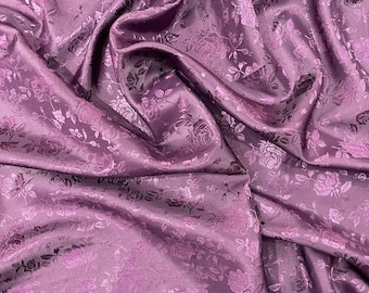 Può includere: Primo piano di un tessuto drappeggiato in una tonalità viola tenue. Il materiale presenta un motivo ripetuto di rose e fogliame in rilievo, creando un aspetto strutturato ed elegante. La lucentezza del tessuto suggerisce una composizione in raso o seta.