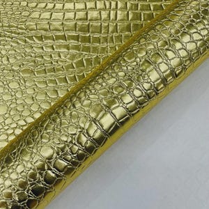 May include: A roll of shiny gold faux leather with a crocodile skin texture. The material has a metallic sheen and a repeating pattern of rectangular and rounded shapes. The material is partially rolled, revealing a white backing.
