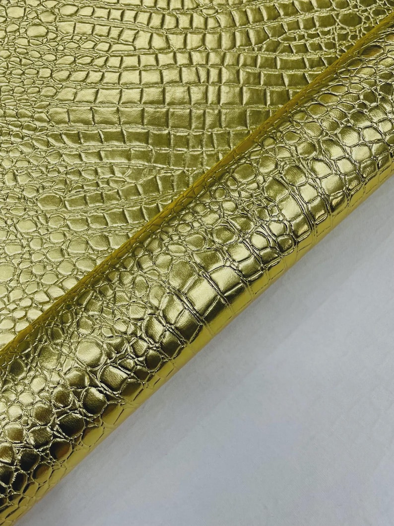 May include: A roll of shiny gold faux leather with a crocodile skin texture. The material has a metallic sheen and a repeating pattern of rectangular and rounded shapes. The material is partially rolled, revealing a white backing.