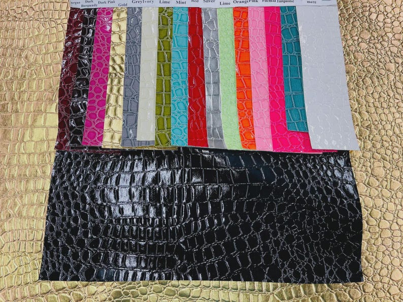 May include: A collection of colorful faux leather swatches, including black, gold, and various shades of pink, green, and blue. The swatches have a crocodile skin texture. The top row has color names printed on it.
