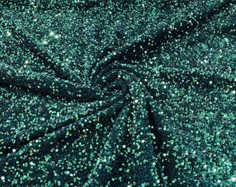 May include: Close-up of a teal sequin fabric. The fabric is densely covered in small, round sequins that catch the light, creating a sparkling effect. The fabric is draped and folded, showing the texture and depth of the material.