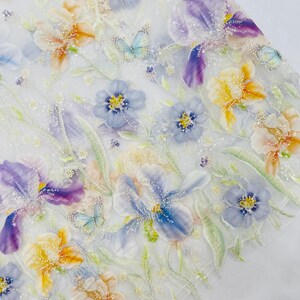 Op de afbeelding: Een doorschijnende witte stof met een complex bloemenpatroon van geborduurde irissen en kleinere bloesems in tinten paars, geel en blauw. Het ontwerp bevat delicate groene bladeren, kleine vlinders en transparante kraalversieringen.