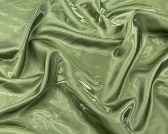 May include: A close-up shot of a piece of olive green fabric with a shiny, satin-like texture. The fabric is draped, creating folds and shadows that highlight its sheen. The material appears lightweight and flowing, suitable for apparel or decorative projects.
