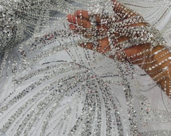 May include: A sheer white fabric featuring an intricate pattern of silver sequins and beads arranged in flowing, curved lines. The material is densely embellished with reflective metallic accents that create a shimmering, textured surface across the mesh.