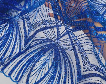 May include: A sheer white fabric featuring an intricate pattern of royal blue sequins and beads arranged in curved, fan-like lines. The material has a scalloped edge and a dense, shimmering texture created by the reflective blue embellishments.