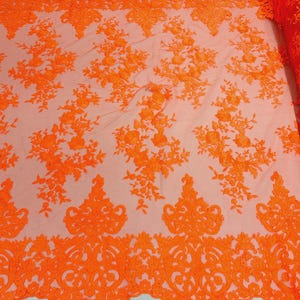 May include: Bright orange lace fabric with floral and ornamental patterns. The fabric is a sheer material with intricate embroidered designs, perfect for apparel or decorative projects. The lace is rolled up on the right side of the image.